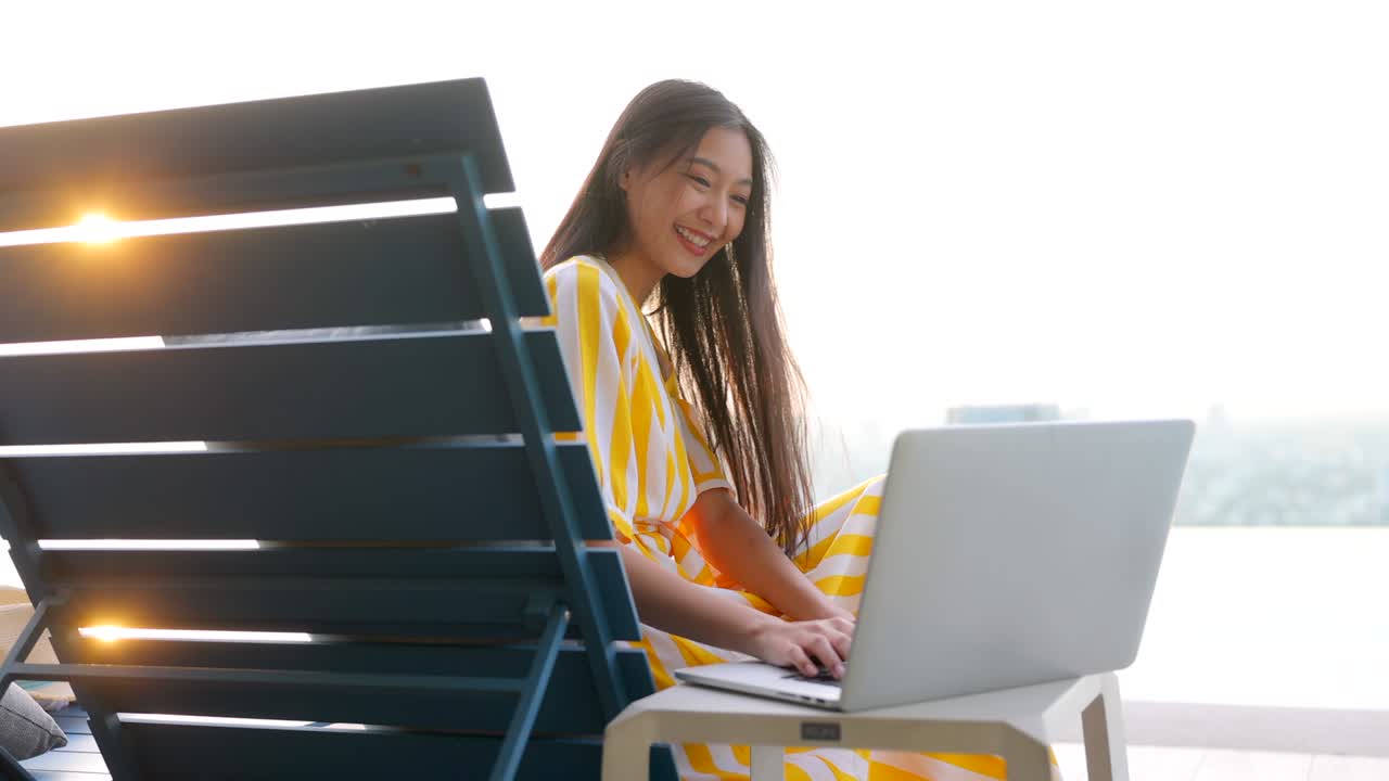 아시안 여성이 수영장  의자에 앉아 편안한 분위기와 밝은 빛을 통해 노트북에 메시지를 입력합니다. 여성은 시내 전망을 즐기고, 해가 지는 자연. 호텔 지붕