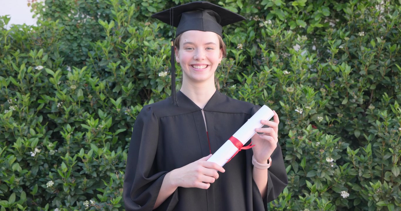 Woman after graduation holding diploma with red ribbon, wearing gown and cap, posing in garden