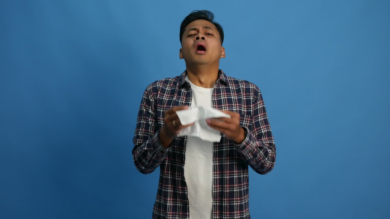 Young Asian man sneezes into tissue. Isolated guy is sick, has a cold or allergic reaction. Health, medicine, illness, treatment concept