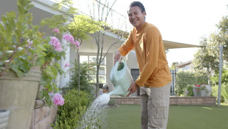 portrait of happy biracial man watering plants in sunny garden, copy space, slow motion