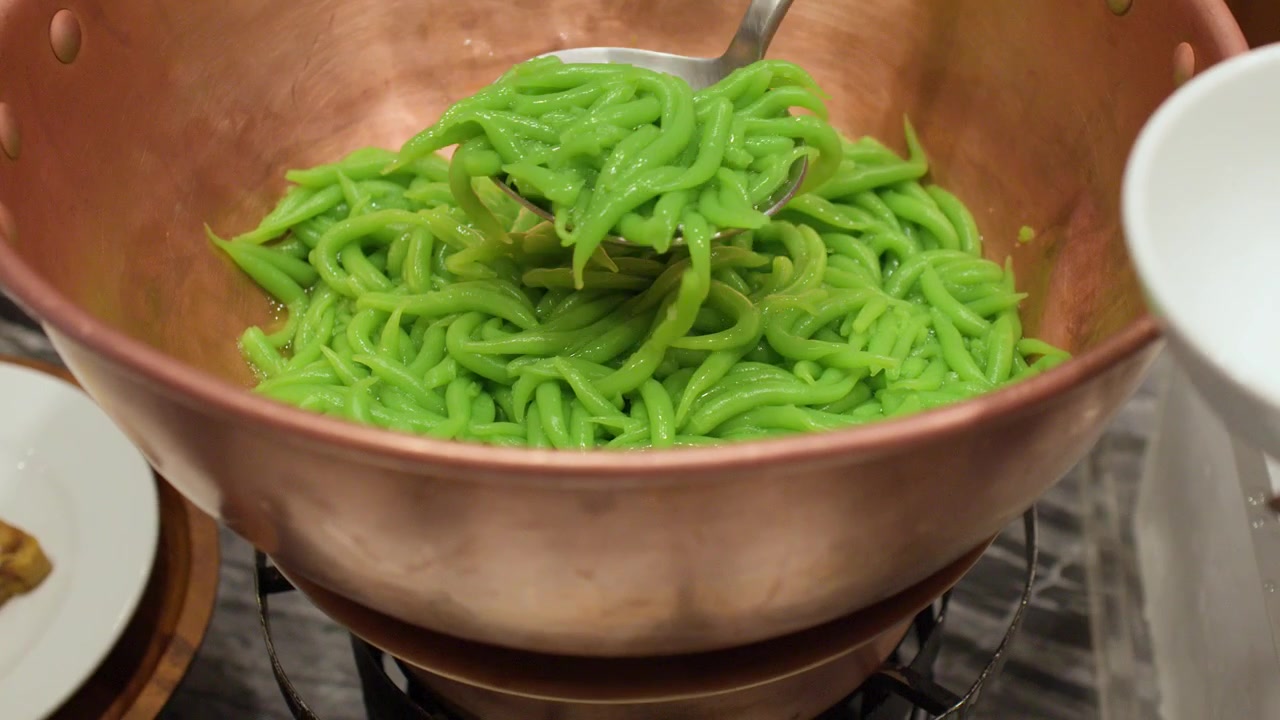 A person scoops green pandan noodles from a copper pot into a white bowl