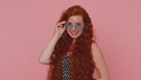 Seductive-cheerful-ginger-stylish-girl-in-dress-wearing-sunglasses,-charming-smile-on-pink-wall