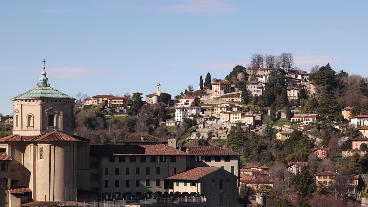 Vista aerea di una storica torre in pietra a Bergamo Alta, Italia, con colline verdi lussureggianti e montagne sullo sfondo.