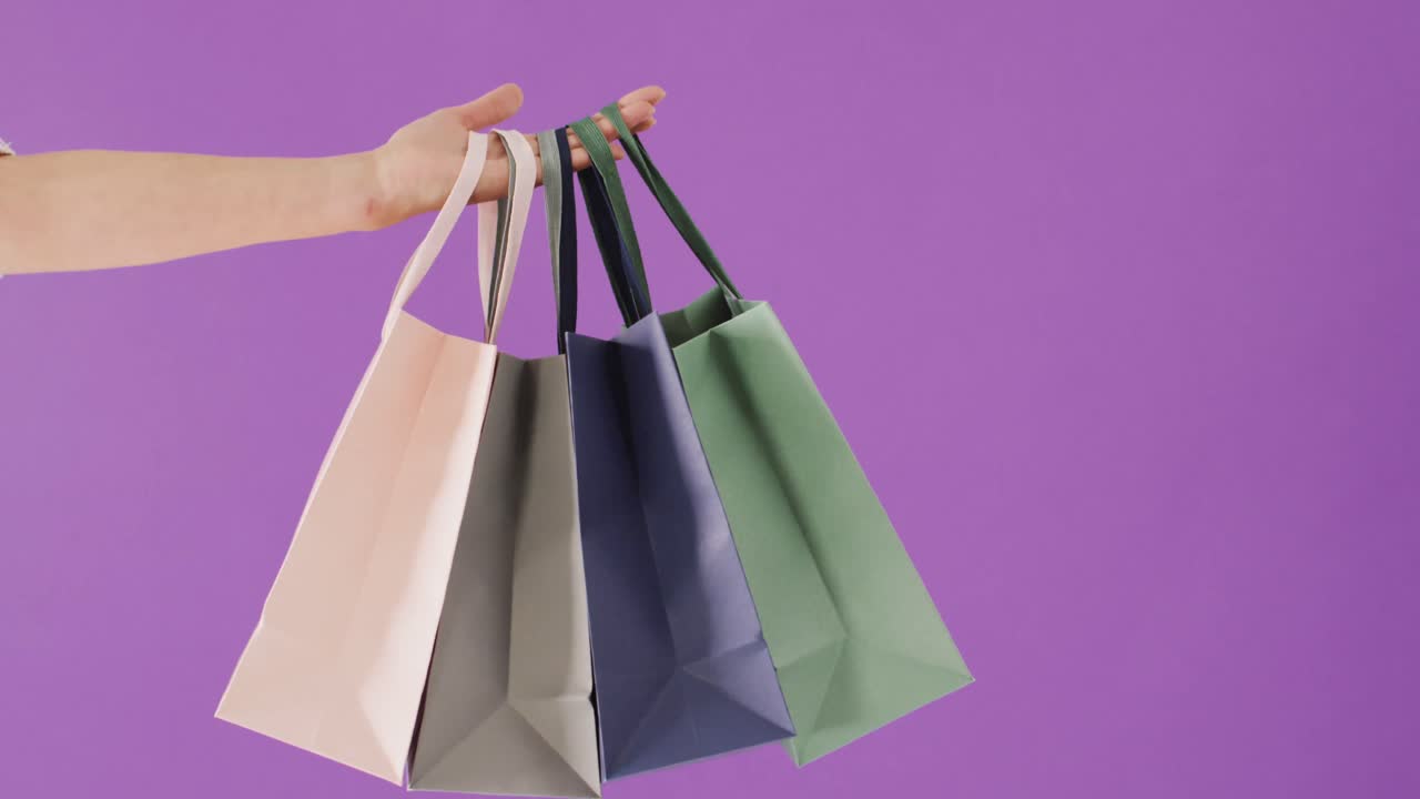 Woman's hand holding coloured gift bags on purple background with copy space