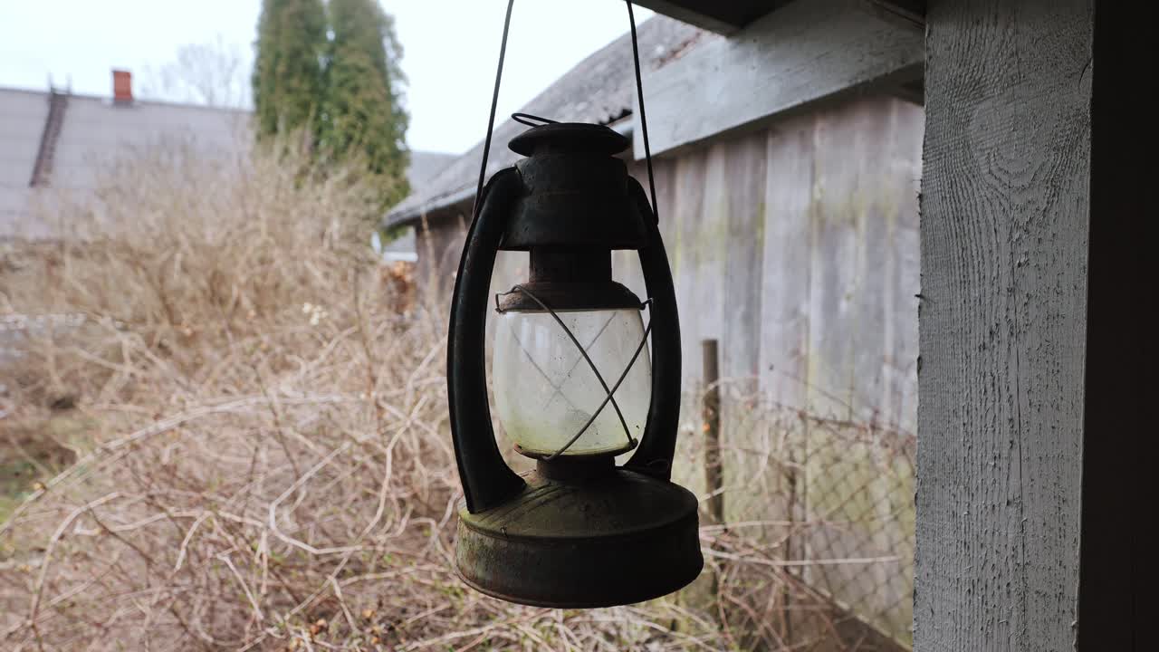 Worn kerosene lantern hangs still near weathered barn in overgrown rural yard
