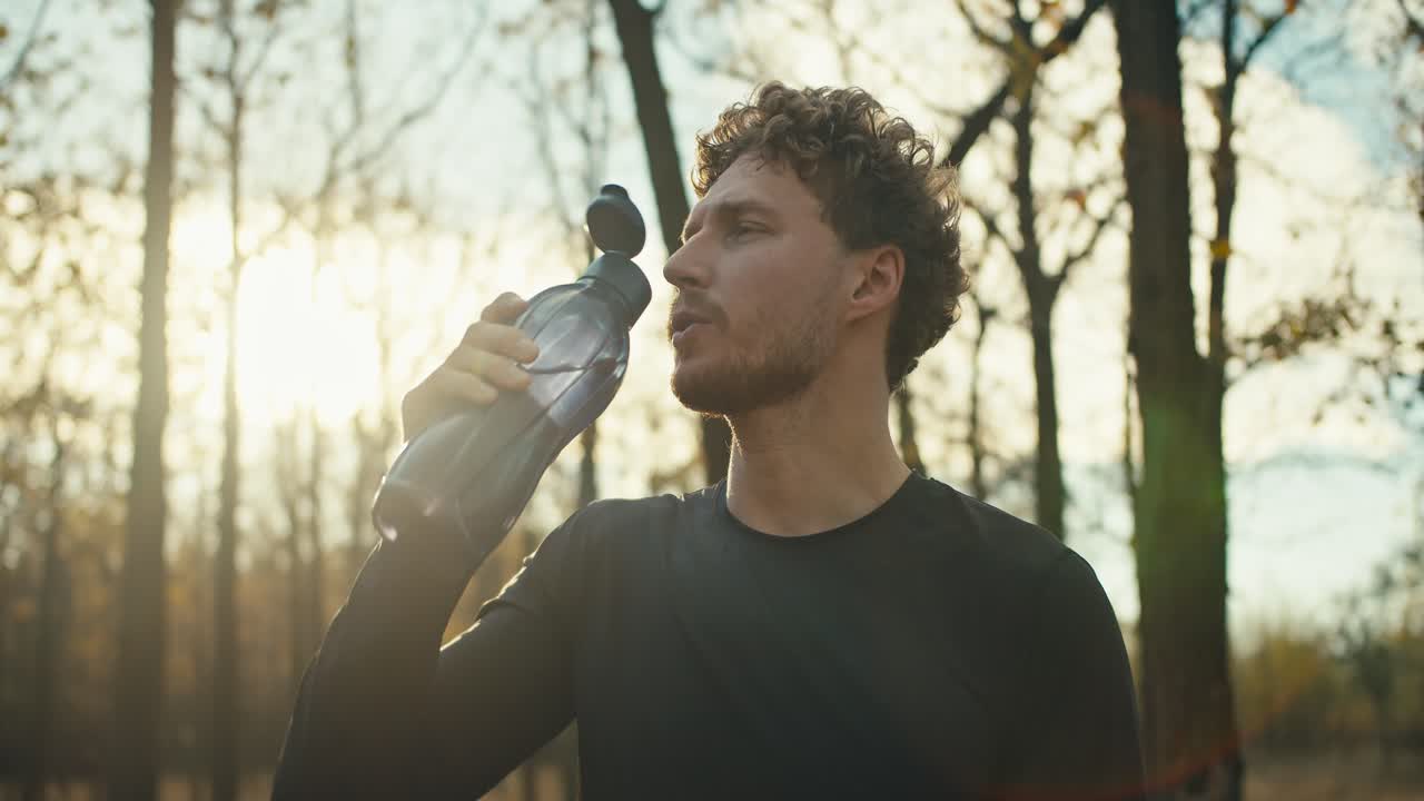atleta masculino confiado con el cabello rizado bebe agua de una botella deportiva gris especial mientras descansa después de una carrera en el bosque de otoño en un día soleado. un hombre en un uniforme deportivo negro bebe agua y descansa luego de jugar deportes y correr