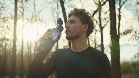 confident male athlete with curly hair drinks water from a special gray sports bottle while resting after a run in the autumn forest on a sunny day. a man in a black sports uniform drinks water and rests after playing sports and jogging