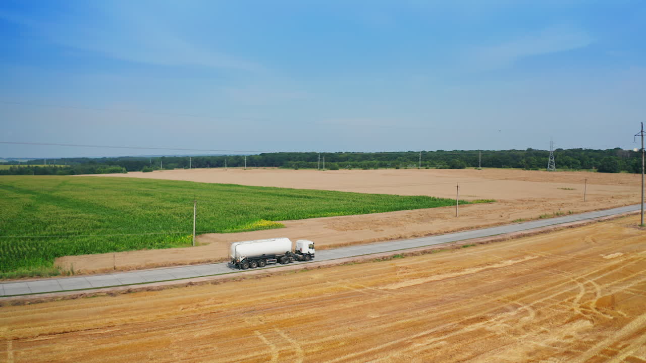 Road crossing the agricultural farmlands and trucks going by it. Tractor loaded with hay bales rides by the cut field. Top view.