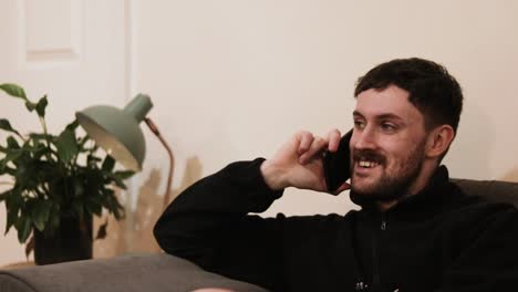 cheerful young man talking on mobile phone sitting on couch in home