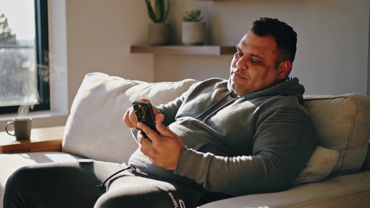 Settling on sofa at home, man in gray hoodie tapping smartphone checking messages near left window