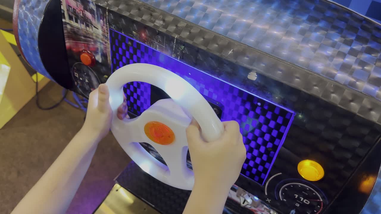 Close-up static shot of a child’s hands gripping a glowing steering wheel on an arcade racing game machine, illuminated with bright blue and orange lights