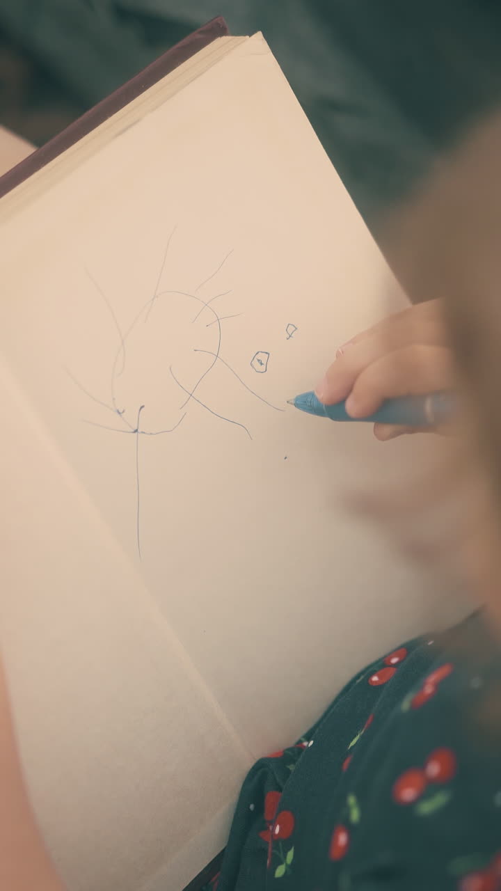 cute little girl draws funny sun in copybook with blue pen sitting on pavement extreme backside close view from above