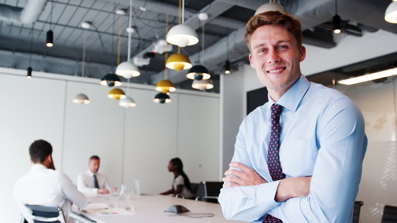 porträt eines geschäftsmanns in einem modernen vorstandssaal mit kollegen, die sich im hintergrund um einen tisch versammeln, in langsamer bewegung.