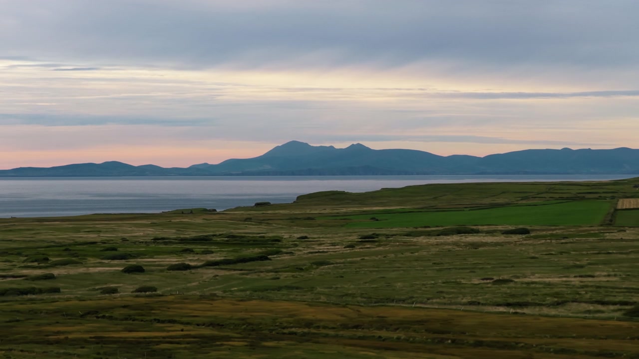 Bølgende grønne åser og gressletter strekker seg over et kystlandskap med fjerne fjell og et rolig hav under en skyet himmel