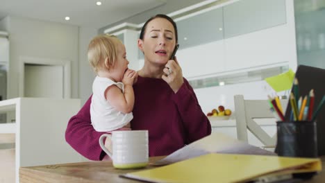 Caucasian-mother-holding-her-baby-and-talking-on-smartphone-while-working-from-home