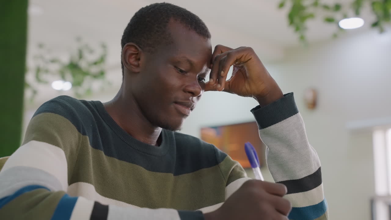 Black man writing love note at table, cozy indoor cafe scene with striped sweater, pen and paper, pensive expression as he drafts message for date and rehearses romantic lines under soft daylight