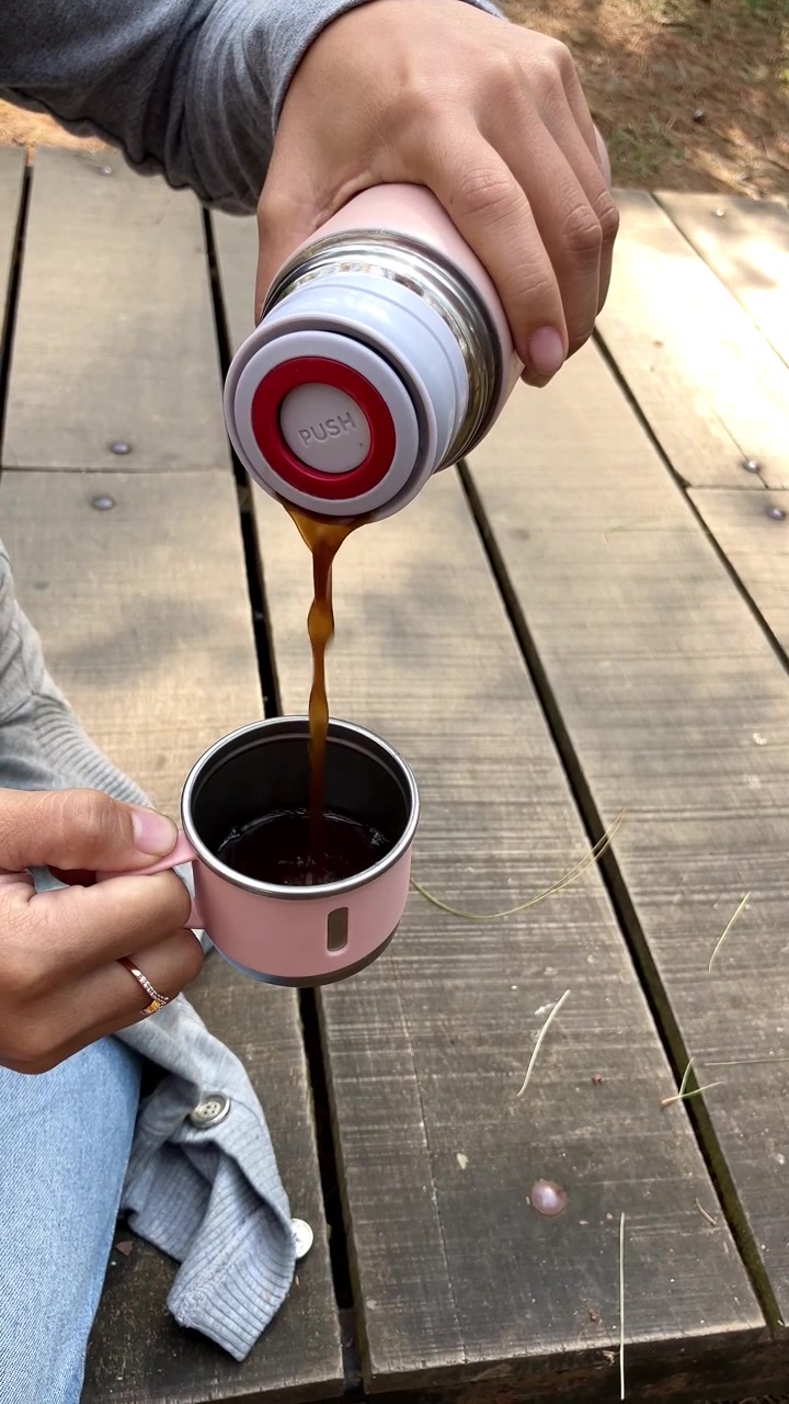 Primer plano a cámara lenta de las manos de una mujer sirviendo una bebida de un termo rosa aislado en una taza a juego. Hermosos pinos altos al fondo. Perfecto para temas de camping o senderismo