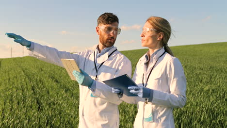 caucasian female and male researchers in white coat, mask and googles using tablet and taking notes while doing pest control in the green field