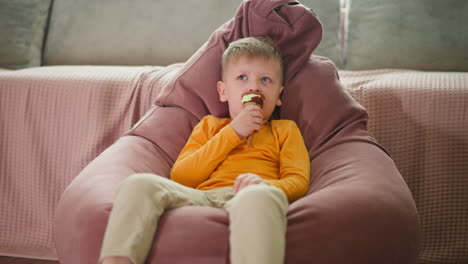 little boy relaxing on pink bean bag chair while enjoying ice cream cone indoors, wearing yellow shirt and light pants, looking upwards thoughtfully