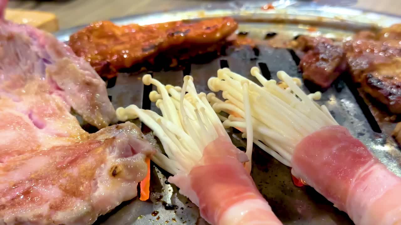 Close-up of enoki mushrooms wrapped in bacon and meat grilling on a hot tabletop grill.