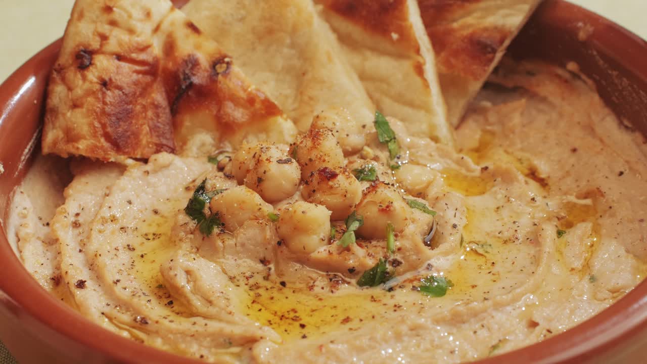 Spreading hummus on bread top view. Hummus garnished with chickpeas, olive oil, spices and greens among fresh vegetables. Humus or fava, a hearty snack of legumes. Kosher healthy vegan food. Lean, vegetarian dish