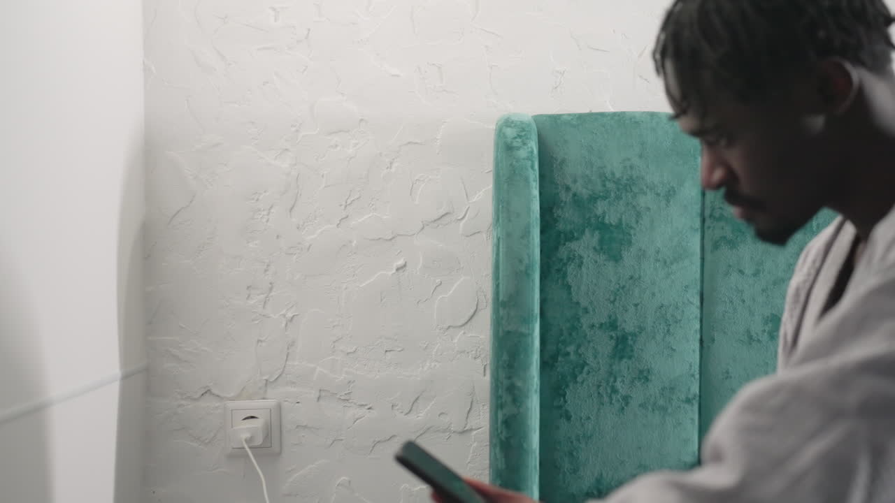 Youth in bathrobe standing by bed while holding phone, as he sit preparing for daily activities. Modern bedroom with teal headboard and white walls, reflecting calm morning atmosphere