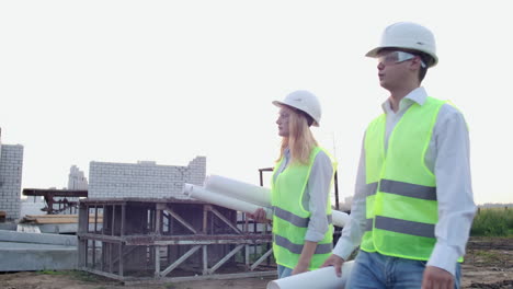hombre y mujer supervisor y asistente con dibujos en cascos blancos van y hablan en el sitio de construcción mostrando la ubicación de los objetos.