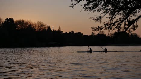 Silueta-De-Dos-Hombres-Remando-En-Canoas-En-La-Presa-Al-Atardecer,-Hora-Dorada