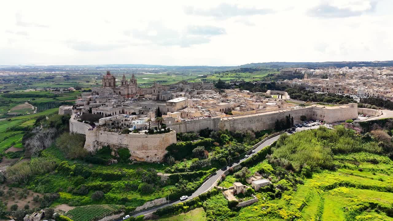 Veduta aerea di Mdina, Malta, che mostra le mura fortificate della città, fitti edifici storici e la campagna verdeggiante con una strada tortuosa sottostante, sotto una chiara luce diurna.