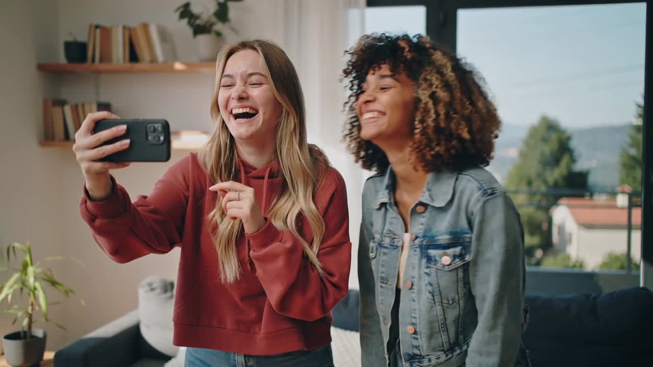Holding smartphone, two women wearing hoodie and denim taking selfie to capture memory at home