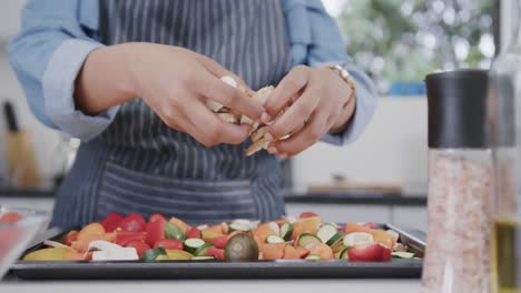 Midsection-of-biracial-woman-in-apron-seasoning-vegetables-on-baking-tray-in-kitchen,-slow-motion