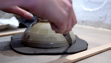 Clay-being-trimmed-in-a-circle-with-knife-using-a-bowl-as-guide-by-hand-on-a-ceramics-workshop,-Handheld-close-up-follow-shot