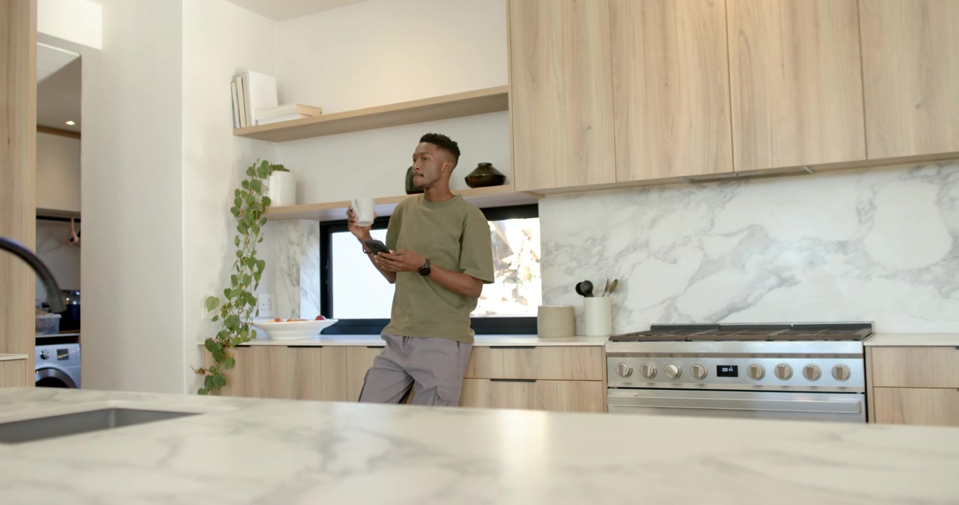 La luce del sole che accarezza un uomo afroamericano che sorseggia da una tazza e legge messaggi sul telefono in cucina, con spazio per il testo.
