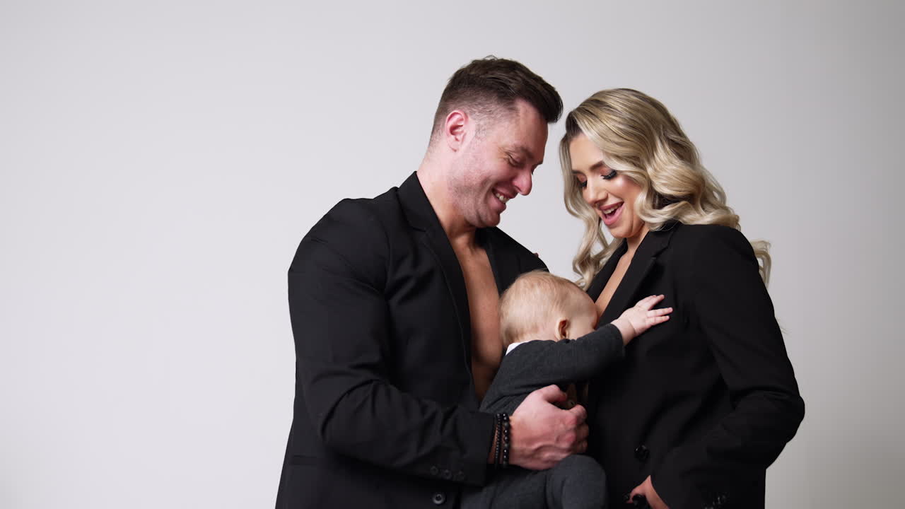 Couple wearing black suits holding a baby. Little child tries to reach mother's breast. Low angle view.