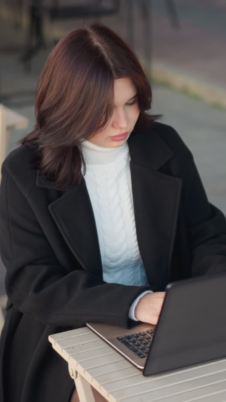 Professional woman working on laptop at an outdoor mall setting, focused and dressed in a stylish black coat, with wooden chairs and tables neatly arranged in the background