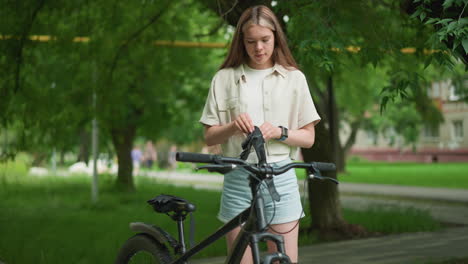 young adult adjusts black gloves beside her bicycle on paved path surrounded by lush greenery, she wears smartwatch on right hand while background shows blurred view of buildings