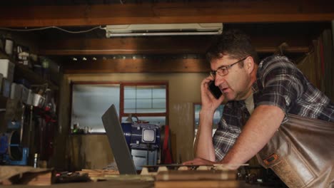 hombre caucásico enfocado en el fabricante de cuchillos en el taller usando portátil y teléfono inteligente