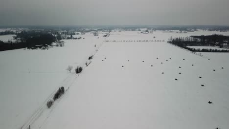 Aerial-flying-over-a-dirt-road-with-power-line,-hay-bales-in-the-field,-winter-landscape