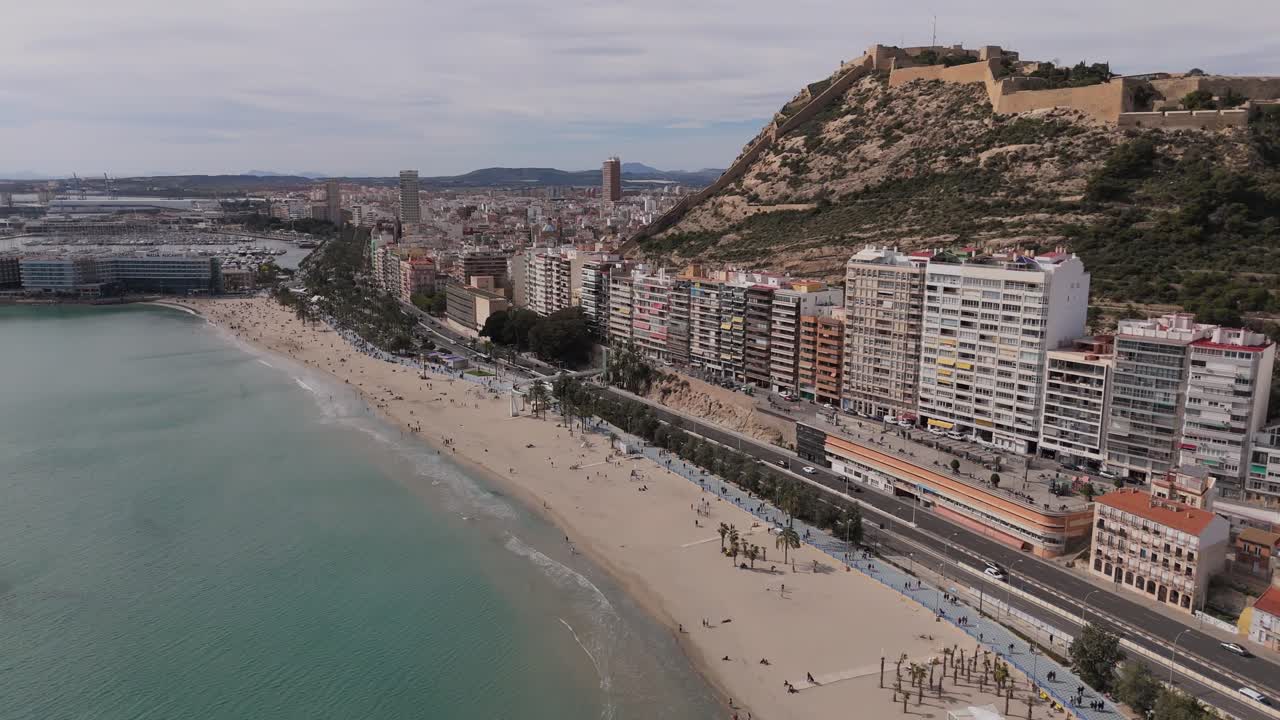 Droneoptagelser der flyver langs Playa del Postiguet i Alicante med byens skyline og Santa Bárbara-slottet på bjerget Benacantil. Middelhavsstrand og promenade på Costa Blanca i Spanien