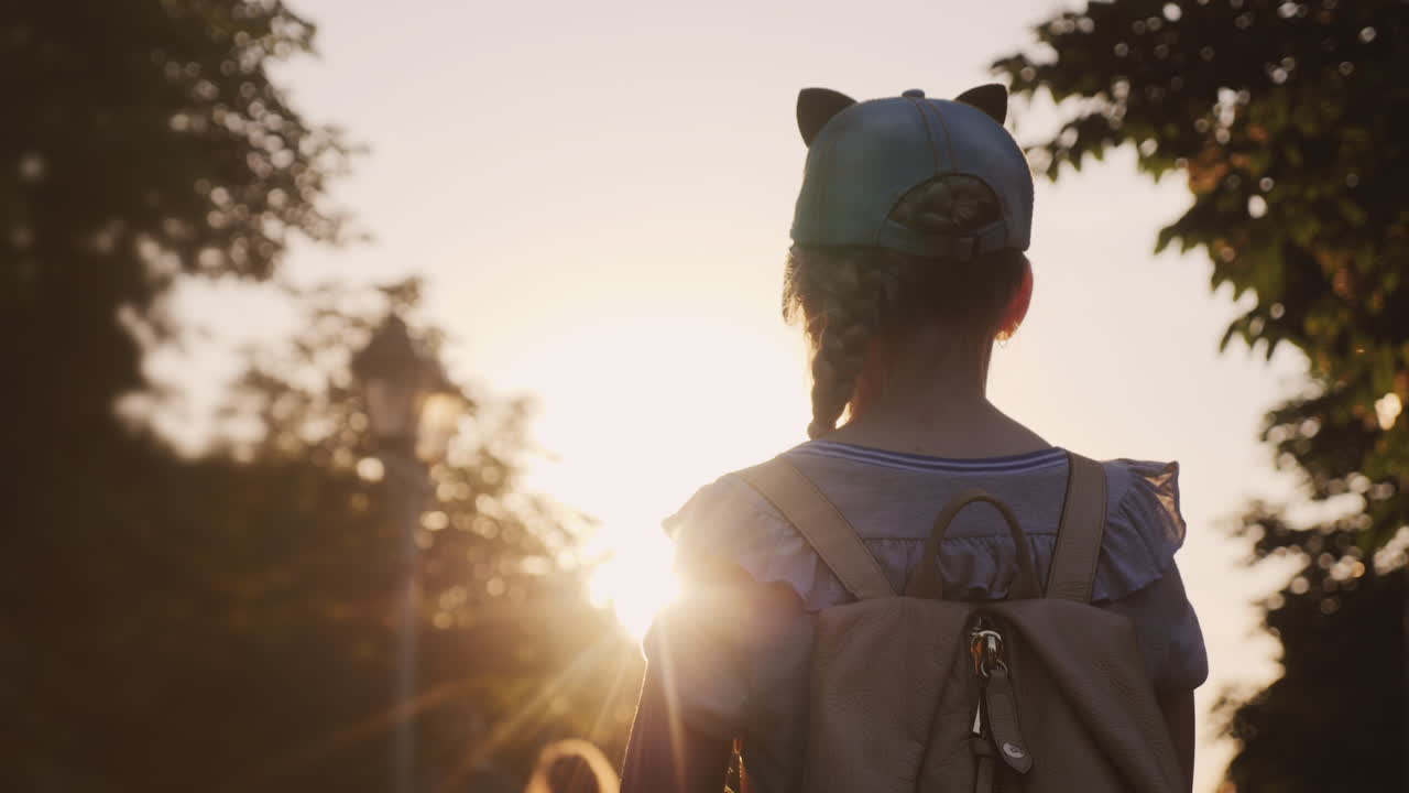 ребенок с рюкзаком стоит в парке, на него светит заходящее солнце