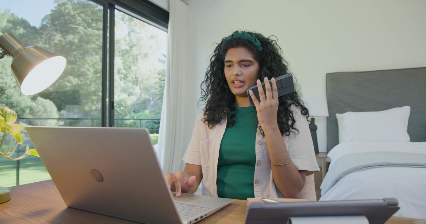 Donna indiana che alza lo smartphone, digitando sul laptop alla scrivania vicino alle porte-finestre, gestendo una chiamata di lavoro in arrivo.