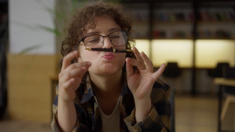 funny girl student with curly hair wearing glasses puts a black pen on her upper lip and laughs while sitting at a table in the library