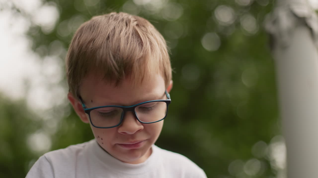 A young boy wearing glasses looks downcast, gazing downward with a sad expression, the background is blurred, showing greenery