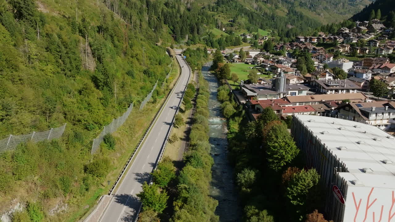 Aerial footage following speedy cars on a highway that runs alongside a river in the Dolomite Mountains.