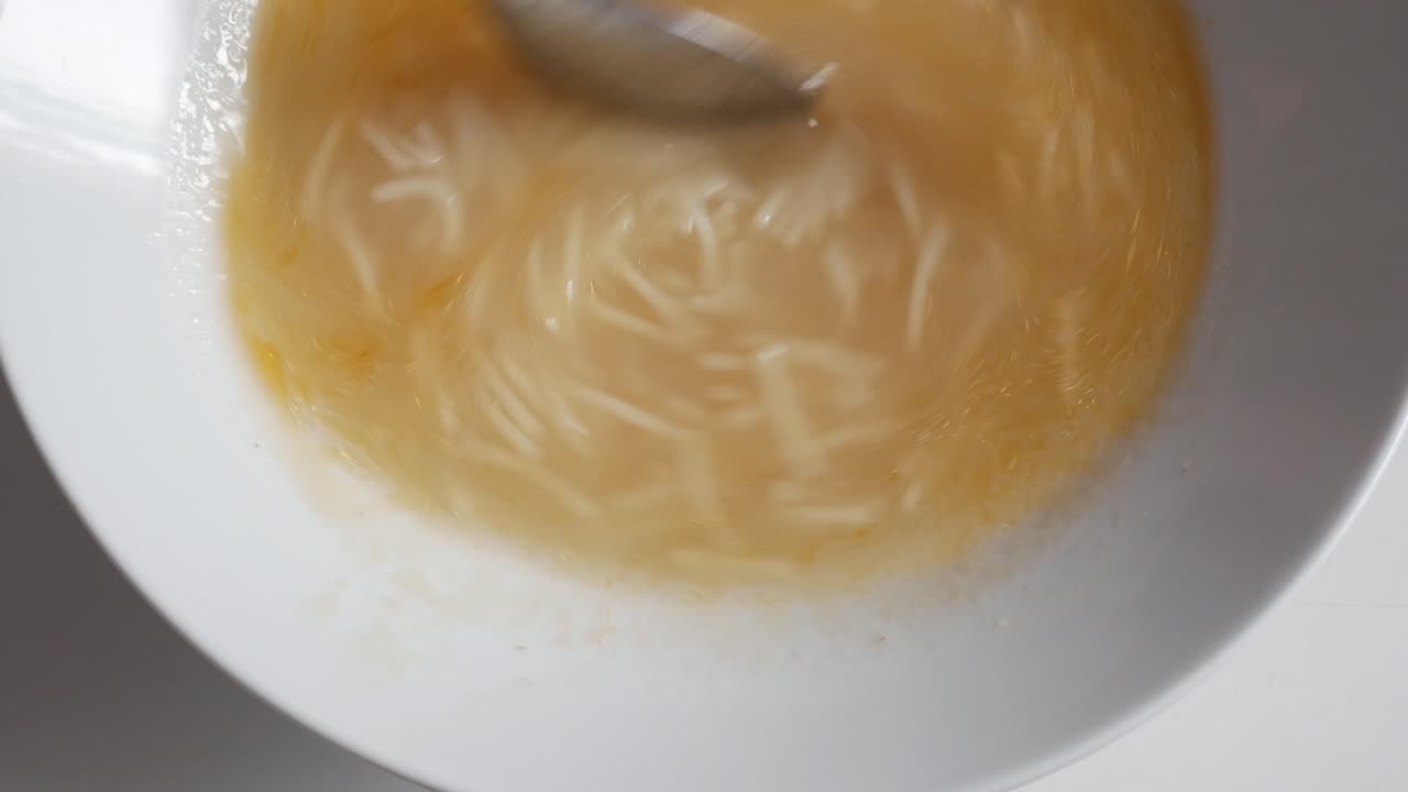 A bowl of cocido soup being stirred with a spoon, with noodles floating in broth