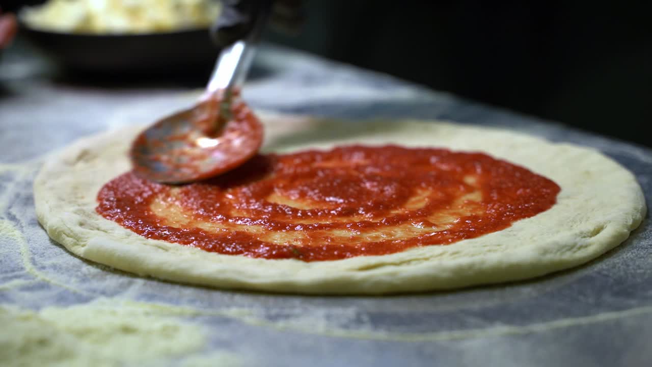 Cucchiaio di metallo che stende la salsa di pomodoro sull'impasto della pizza.