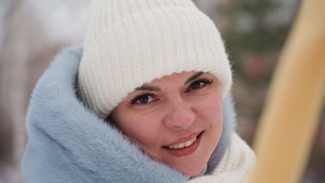 Ritratto ravvicinato di mamma sorridente con cappotto invernale, berretto bianco e sciarpa blu con cappuccio all'aperto, sfondo bokeh morbido, luce naturale che evidenzia le guance e un'espressione calda, atmosfera accogliente in un parco innevato.