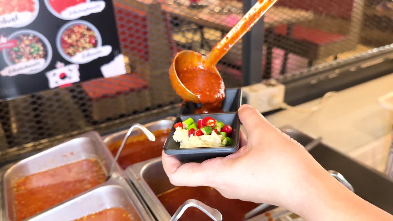 A person ladles chili garlic sauce into a small dish at a Korean BBQ stall in Bangkok, Thailand. Bright lighting enhances the vibrant colors