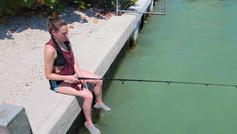 mujer pescando mientras se sienta en el borde del muelle en la orilla mientras enrolla la caña giratoria y sonríe a la cámara