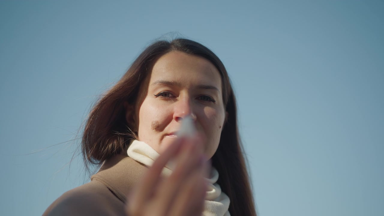 Primo piano di una donna all'aperto contro un cielo azzurro limpido, che indossa una sciarpa di colore chiaro e un cappotto marrone. I capelli si muovono leggermente nel vento mentre la donna gesticola con la mano tenendo un piccolo oggetto bianco. Illuminazione naturale soffusa